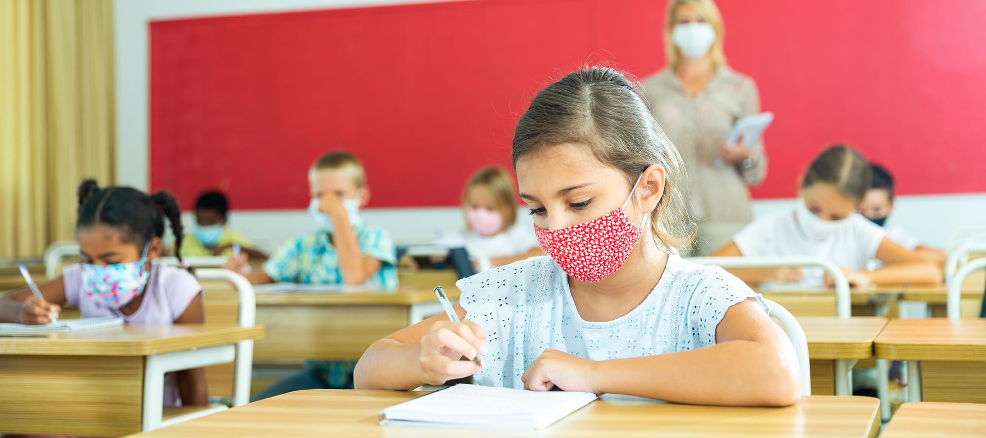 Child with medical mask sitting in classroom with classmates and teacher
