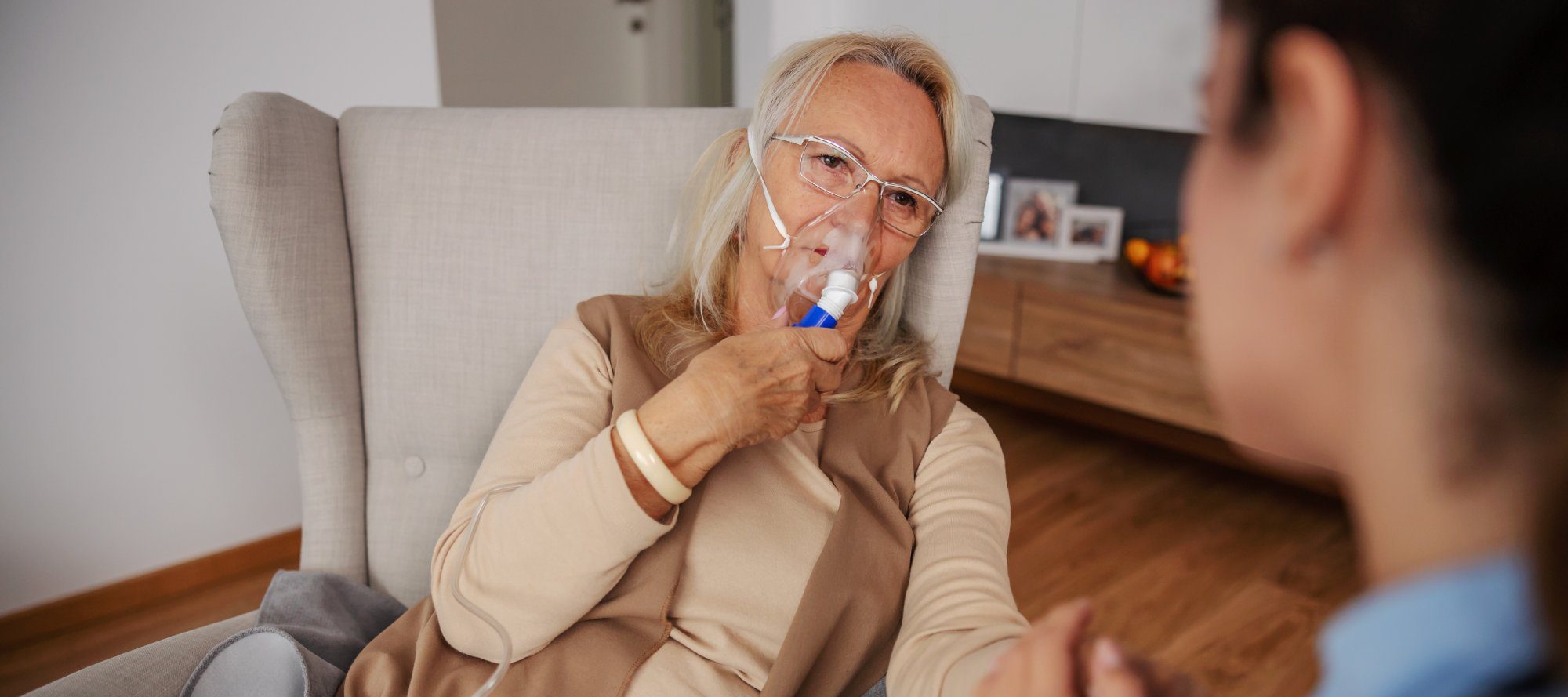 Ill blond senior woman sitting at home, taking oxygen from respirator while nurse sitting next to her and holding her hand.
