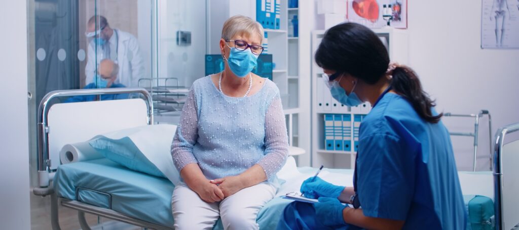 Nurse in protective equipment questioning senior lady patient who wears a mask.