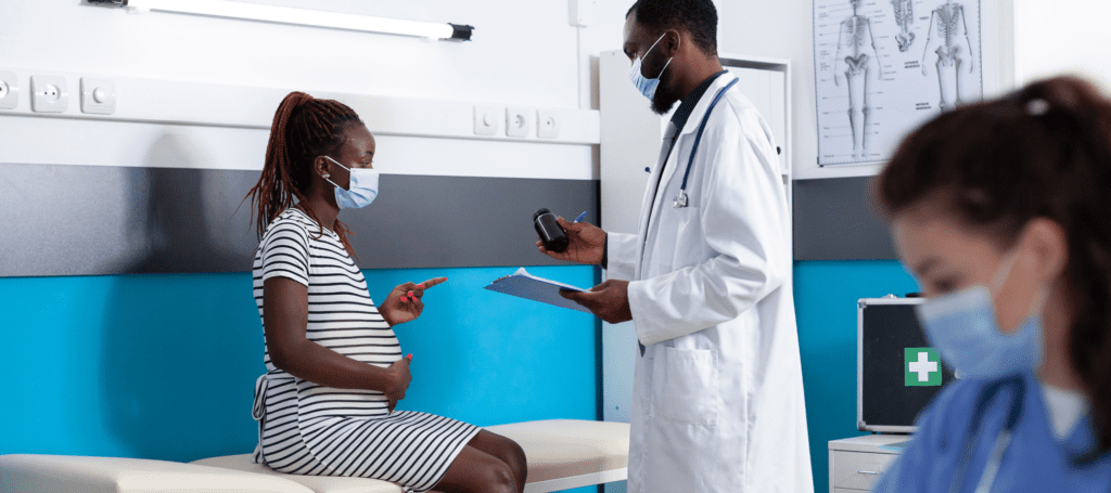 Pregnant woman sitting on medical exam table speaking with her doctor both wearing masks