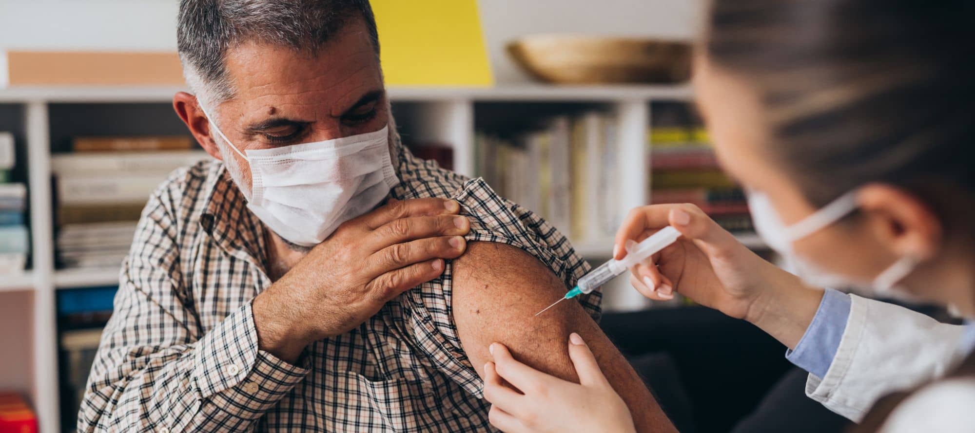 Mature man holding his shirt sleeve up while wearing mask as doctor administers vaccine in upper arm