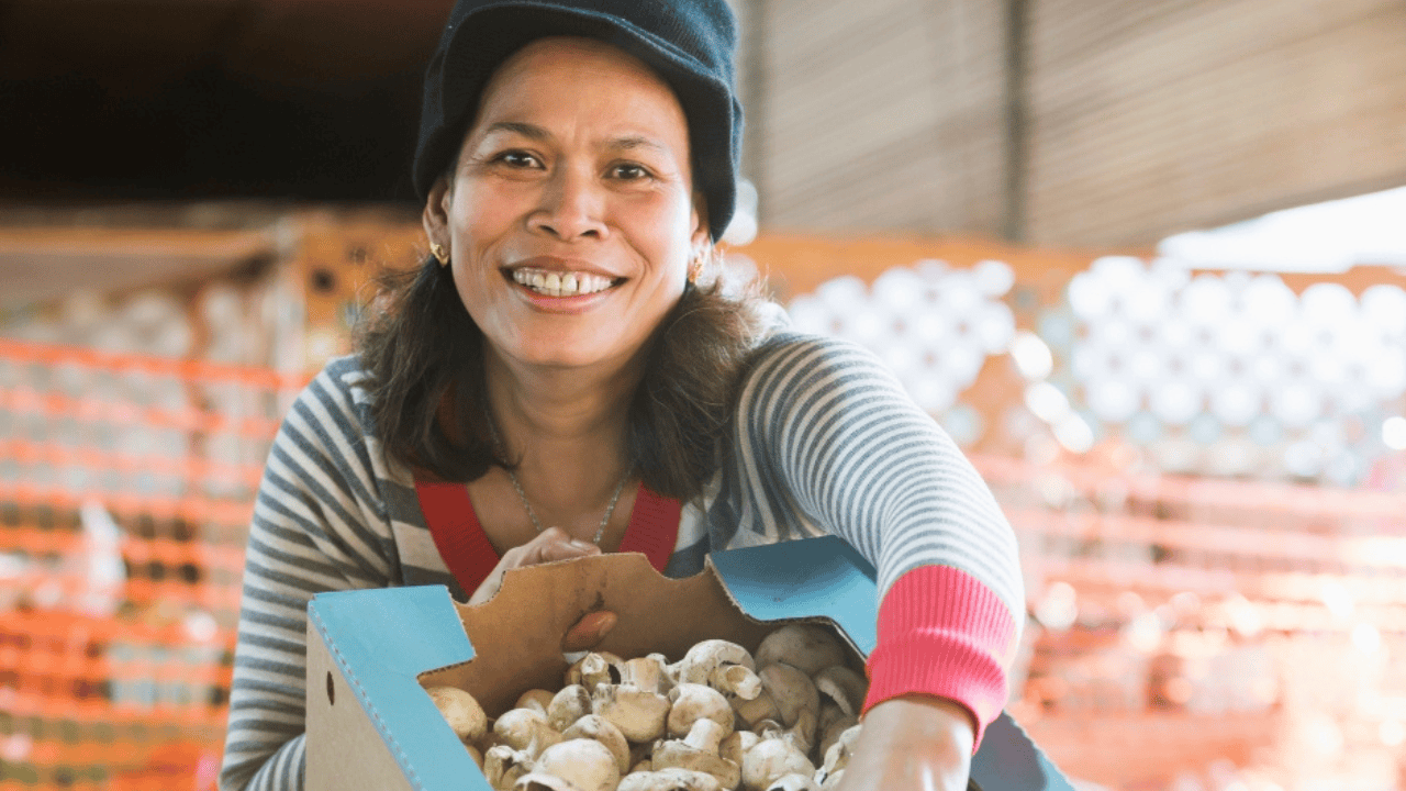 A middle aged woman smiling as she holds a cardboard box of button mushrooms