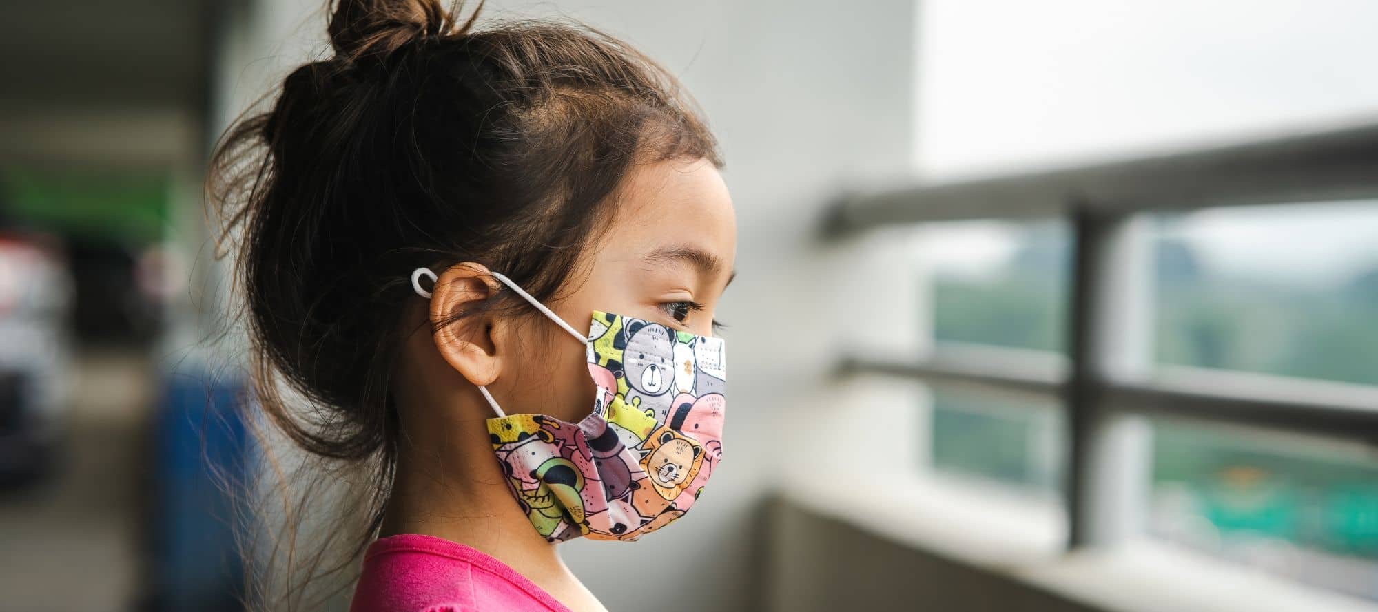 Young child wearing mask with animals on it looking outside of a window