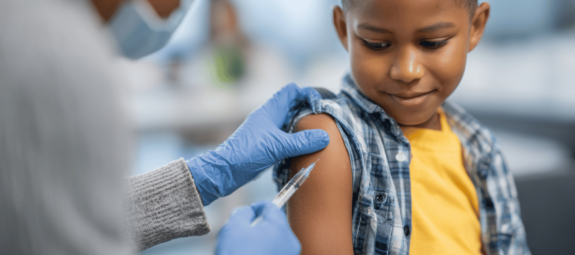 a young black child wearing a plaid sweater and yellow shirt receiving a shot